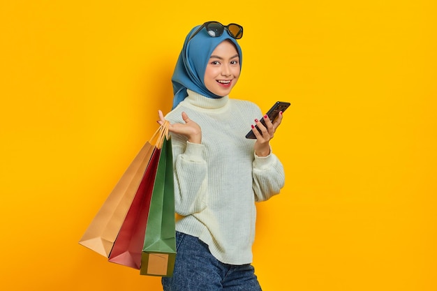 Bella donna asiatica allegra in maglione bianco che tiene borse della spesa utilizzando il telefono cellulare guardando la fotocamera isolata su sfondo giallo
