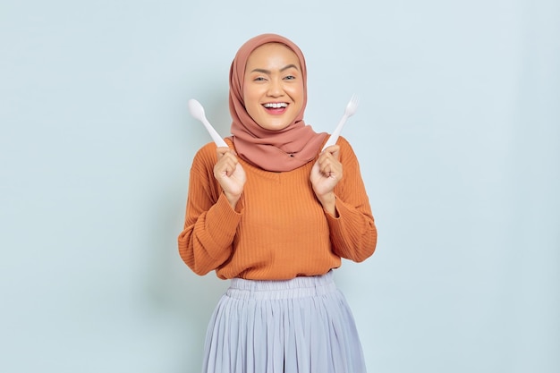 Bella donna asiatica allegra in maglione marrone e hijab tenere forchetta e cucchiaio prima di colazione isolato su sfondo bianco