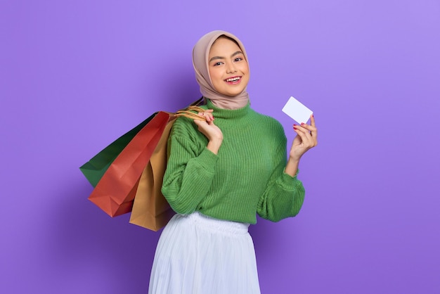 Bella donna asiatica allegra in maglione verde che tiene borse della spesa e carta di credito isolate su sfondo viola