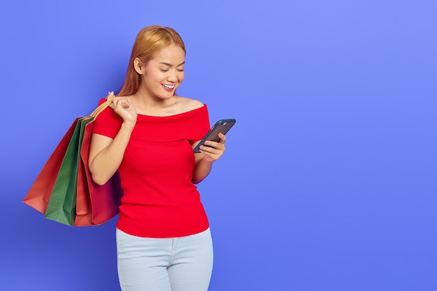 Bella donna asiatica allegra in vestito rosso che tiene borse della spesa, utilizzando il telefono cellulare isolato su sfondo viola