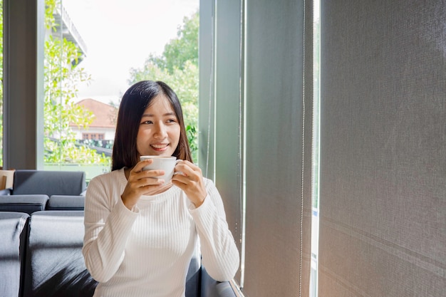 Bella donna asiatica beve una tazza di caffè e tè in soggiorno con finestra a tendina