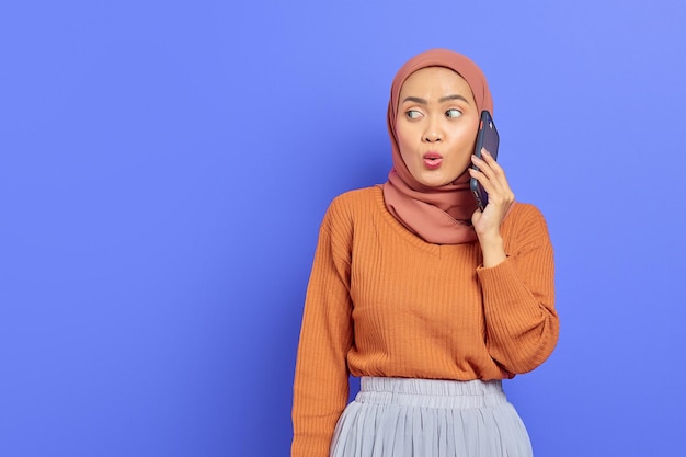 Bella donna asiatica carina in maglione marrone e hijab che parla sul cellulare mentre guarda lateralmente isolato su sfondo viola. La gente islam concetto religioso