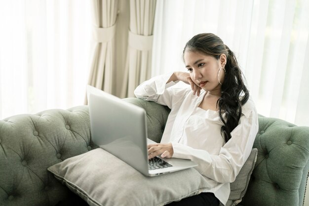 Bella donna asiatica casuale felice che lavora ad un computer portatile che si siede sullo strato in casa.