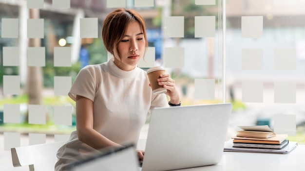 Bella donna asiatica che tiene la tazza da caffè seduta sul computer portatile funzionante nell'ufficio moderno.