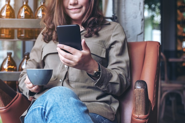 bella donna asiatica che tiene, utilizza e guarda smart phone mentre beve il caffè nella caffetteria