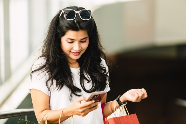 Bella donna asiatica che usa lo smartphone e tiene le borse della spesa nel centro commerciale