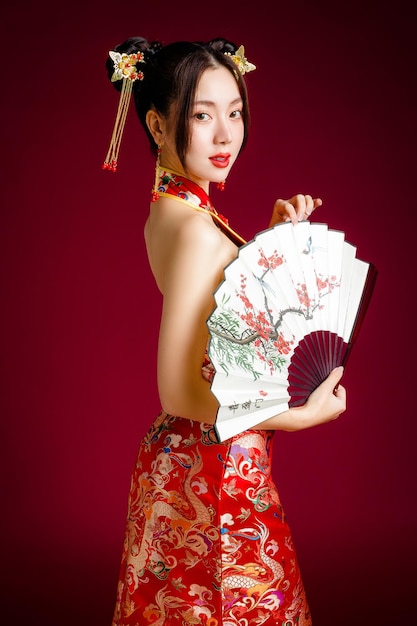 Bella donna asiatica con la pelle pulita e fresca che indossa un abito tradizionale cinese che tiene il ventaglio in posa su sfondo rosso Il testo cinese significa cercare la primavera e felice di vedere la prugna