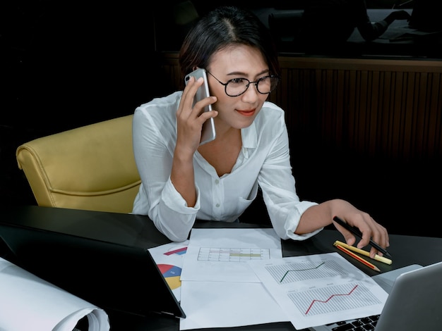 Bella donna asiatica felice che parla con il telefono cellulare mentre si lavora con il computer portatile in ufficio