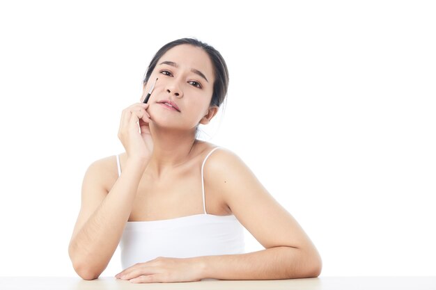 Bella donna asiatica guardando la telecamera e con un piccolo pennello cosmetico in lei ha sollevato una mano per modellare il suo colpo alla testa del sopracciglio con le spalle nude sulla superficie bianca