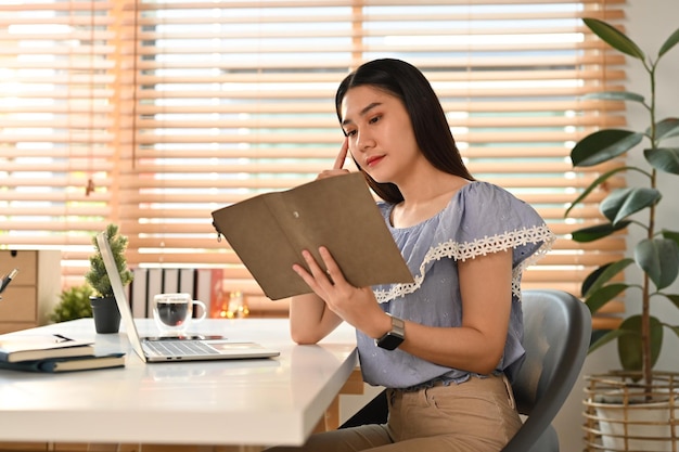 Bella donna asiatica imprenditrice seduta in un posto di lavoro confortevole e leggendo le note nel pianificatore