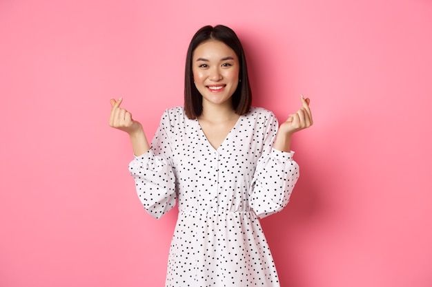 Bella donna asiatica in abito che mostra segni di cuore coreano e sorridente, in piedi su sfondo rosa romantico.