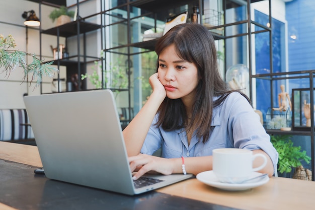 Bella donna asiatica in camicia blu facendo uso del computer portatile e bevendo caffè