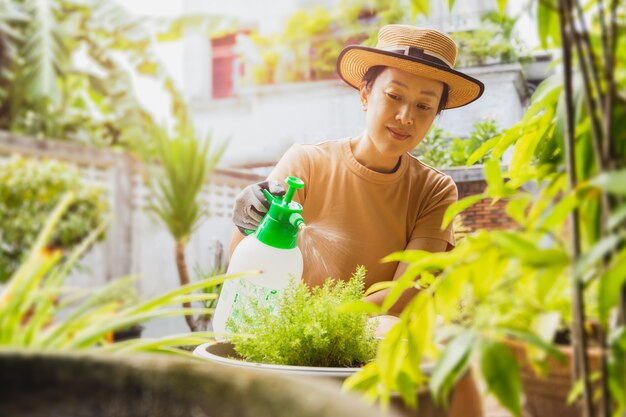 Bella donna asiatica in cappello che innaffia la pianta da una bottiglia spray nebbiosa in giardino