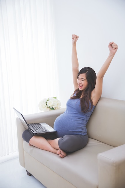 Bella donna asiatica incinta che si siede sul divano con un computer portatile