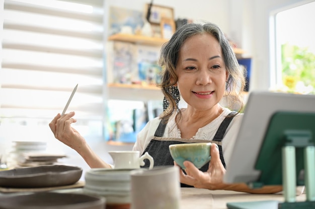 Bella donna asiatica invecchiata che impara una lezione di pittura su ceramica online tramite tablet nel suo studio