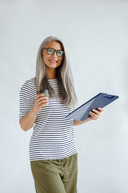 Bella donna asiatica matura in abiti casual tiene appunti e tazza in piedi su sfondo grigio chiaro in studio