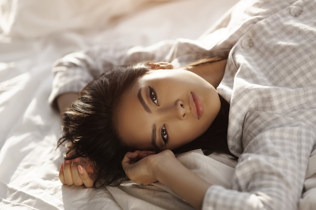 Bella donna asiatica sdraiata sul letto nella sua stanza a casa. Ragazza calma in camicia da notte con capelli lunghi che si rilassano a letto nella mattina soleggiata.