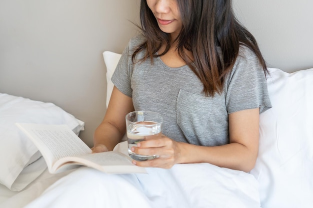 Bella donna asiatica seduta a letto a leggere un libro mentre si tiene un bicchiere di acqua minerale al mattino a casa Relax e stile di vita nel concetto di giorno libero Primo piano