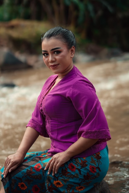 Bella donna asiatica seduta sulla roccia vicino al fiume con un volto sorridente mentre indossa un abito viola