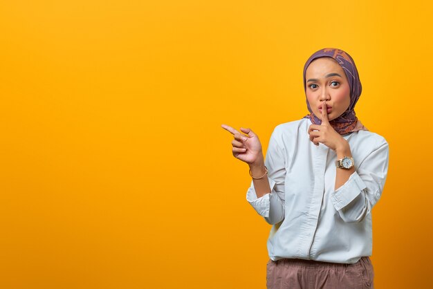 Bella donna asiatica segnale di tacere e indicando il lato su sfondo giallo