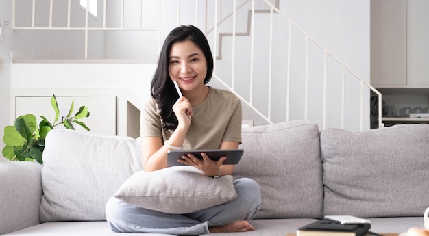 Bella donna asiatica sorridente che guarda l'obbiettivo rilassante sul divano in pelle a casa di lavoro su un computer portatile