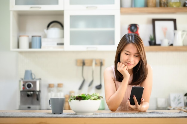 Bella donna asiatica sorridente che utilizza il cellulare dello smartphone in cucina