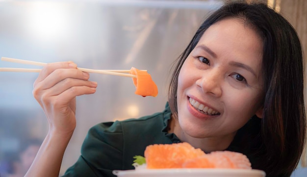 Bella donna asiatica sorridente e felicemente guardando la telecamera che mangia sashimi di salmone