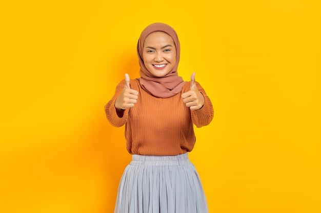 Bella donna asiatica sorridente in maglione marrone e hijab che mostra i pollici in su gesto con la mano isolata su sfondo giallo. La gente islam concetto religioso