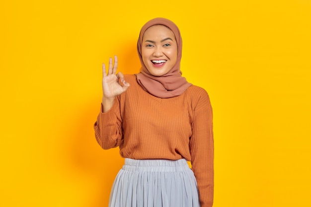 Bella donna asiatica sorridente in maglione marrone e hijab che mostra un gesto ok dimostra il simbolo di approvazione isolato su sfondo giallo. La gente islam concetto religioso