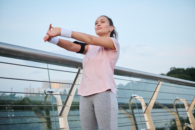 Bella donna, atleta che indossa una maglietta rosa allunga il suo corpo, in piedi su un ponte di vetro della città durante l'allenamento mattutino all'alba. Sport, stile di vita sano e attivo, vitalità, concetto di perdita di peso