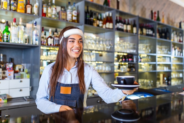 Bella donna barista è in possesso di una tazza di caffè caldo, indossa una visiera mentre si trovava vicino al bancone del bar nella caffetteria. Nuovo normale