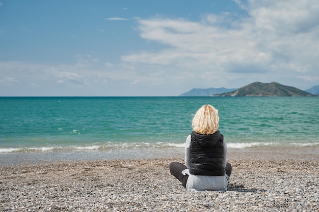 Bella donna bionda adulta in abbigliamento sportivo si siede su una spiaggia di ciottoli vicino all'azzurro del mare Egeo luce solare naturale mezzogiorno Vista dal retro sullo sfondo del mare e onde vista posteriore