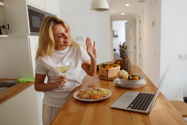 Bella donna bionda che agita la mano e video chat sul laptop Pranzo a casa con vino