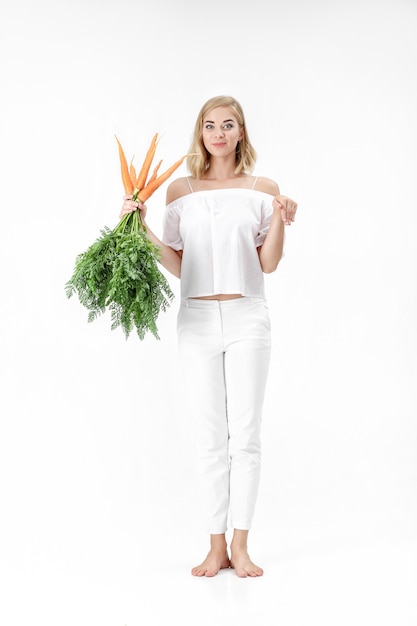 Bella donna bionda che mostra il coniglio e che tiene una carota fresca con foglie verdi su sfondo bianco. Salute e dieta