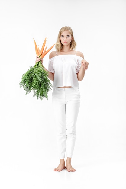 Bella donna bionda che mostra il coniglio e che tiene una carota fresca con foglie verdi su sfondo bianco. Salute e dieta