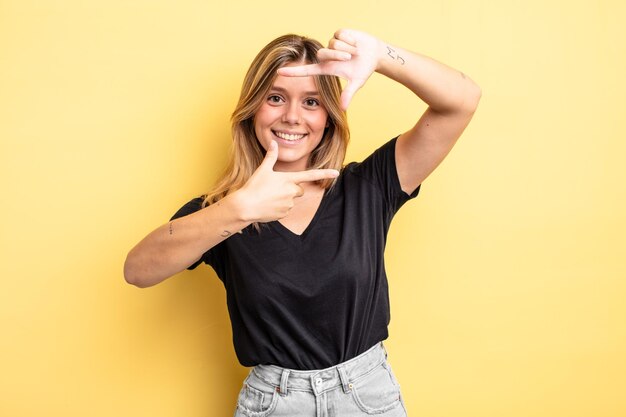 Bella donna bionda che si sente felice, amichevole e positiva, sorride e fa un ritratto o una cornice per foto con le mani