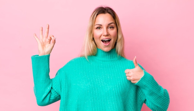 Bella donna bionda che si sente felice stupita soddisfatta e sorpresa che mostra bene e pollice in alto gesti sorridenti