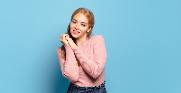 Bella donna bionda che si sente innamorata e sembra carina, adorabile e felice, sorridendo romanticamente con le mani accanto al viso