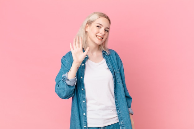 Bella donna bionda che sorride felicemente, agitando la mano, dando il benvenuto e salutandoti