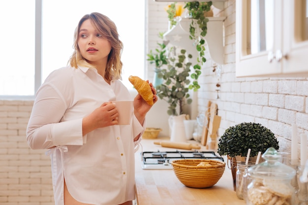 Bella donna bionda che tiene in mano un gustoso croissant mentre è in piedi in cucina