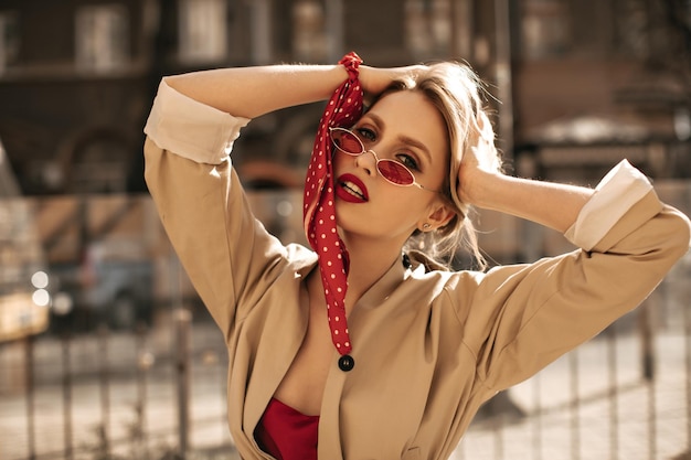 Bella donna bionda con labbra rosse in occhiali da sole colorati e trench beige guarda nella fotocamera e cammina all'aperto