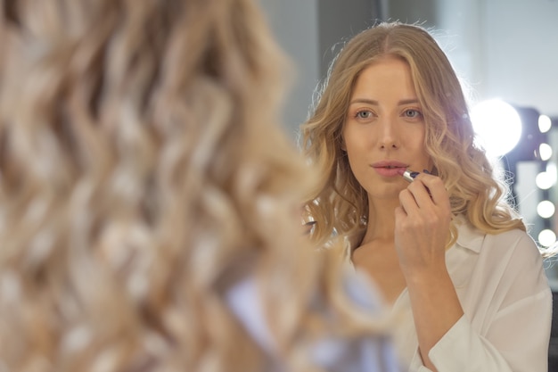 Bella donna bionda con una bella acconciatura che guarda il suo riflesso nello specchio e dipinge le labbra al salone di bellezza