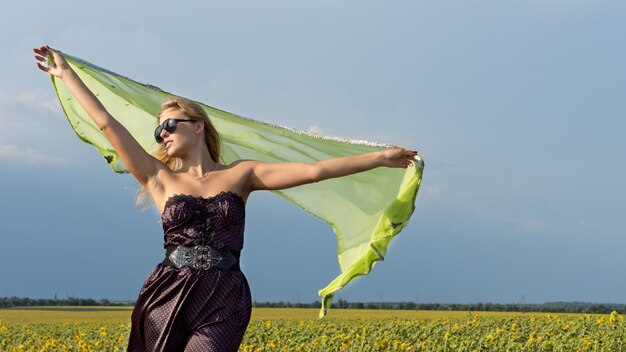 Bella donna bionda con una sciarpa tenuta alta nell'aria che soffia nel vento in piedi in aperta campagna con un campo di girasoli