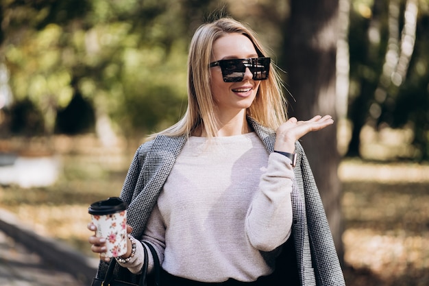 Bella donna bionda con una tazza di caffè sulla strada. Ritratto di donna d'affari sorridente alla moda in pantaloni scuri, maglione cremoso e occhiali da sole. Concetto di moda. Stile d'affari femminile. Alta risoluzione.