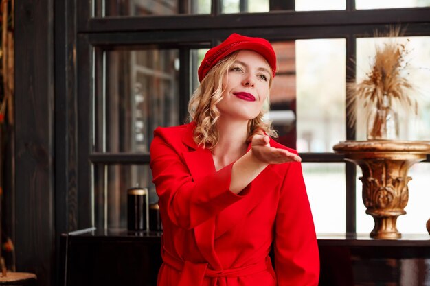 Bella donna bionda elegante in berretto rosso e giacca rossa che soffia un bacio