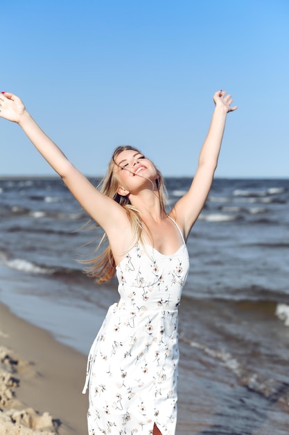 Bella donna bionda felice sulla spiaggia dell'oceano in piedi in un abito estivo bianco, alzando le mani