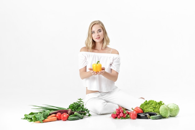 Bella donna bionda in abiti bianchi e un sacco di verdure fresche su uno sfondo bianco. La ragazza tiene il peperone dolce