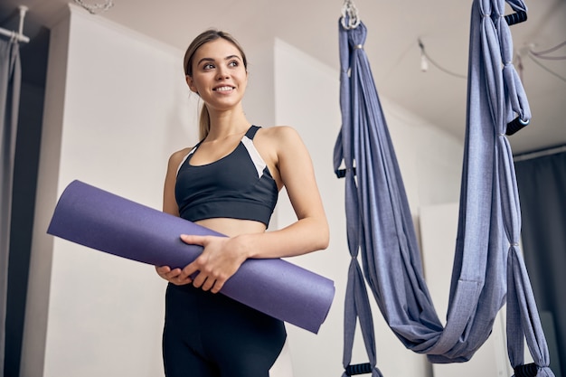 Bella donna bionda in abiti sportivi in piedi in palestra vicino alle amache mentre tiene in mano un tappetino da yoga