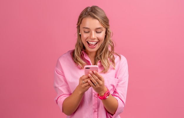 Bella donna bionda in camicia rosa sorridente che tiene tenendo utilizzando lo smartphone in posa su sfondo rosa isolato sorridente divertendosi emozionato eccitato