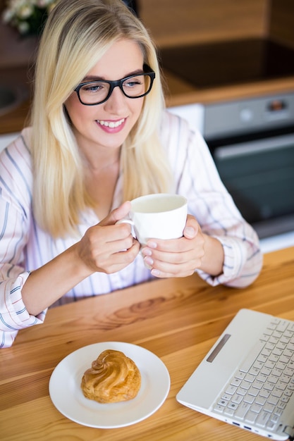 Bella donna bionda in pigiama che beve caffè e usa il portatile a casa